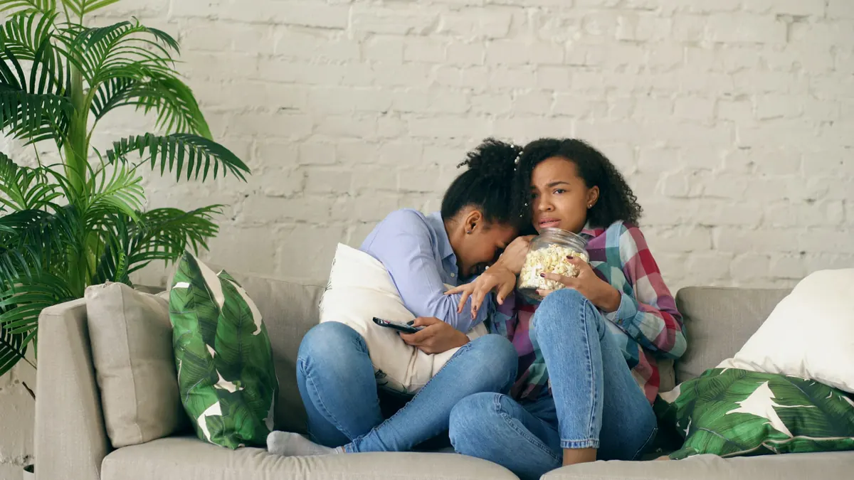 Two friends on a sofa sharing popcorn while watching TV at home